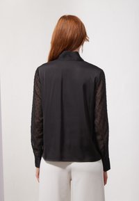 Black blouse with a silk front and sheer, dotted sleeves. Features a collar and button closure. Tailored fit with a smooth texture.