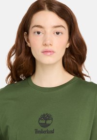 Olivgrön bomulls t-shirt med rund hals, med en svart Timberland-logotypgrafik i mitten. Slät textur, avslappnad design.