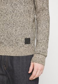 Pull en maille marron clair avec une texture mouchetée, présentant un patch logo noir à l'ourlet et des poignets côtelés. Porté sur un jean foncé.