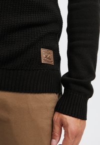 Schwarzer gestrickter Pullover mit geripptem Saum und Bündchen, der einen strukturierten Körper hat. Ein braunes Lederpatch mit "Solid"-Branding befindet sich an der Seite.