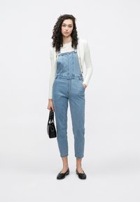 Lys blå denimloffer med rett snitt, justerbare stropper og frontlommer på forstykket. Brukt med en hvit topp og svarte flate sko.