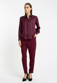 Bordowa satynowa bluzka z guzikami i długimi rękawami w połączeniu z dopasowanymi spodniami. Czarnym szpilkom dopełnia stylizację.