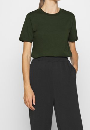 Mujer con camiseta de manga corta y cuello redondo de color verde oscuro metida dentro de pantalones negros de cintura alta, con una mano en el bolsillo.