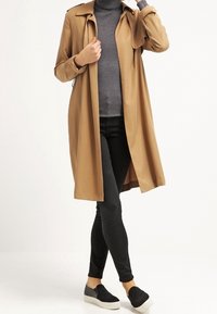 Kobieta w długim, camelowym płaszczu na grubej, szarej golfie, czarnych, dopasowanych spodniach i czarnych butach wsuwanych z białymi podeszwami.