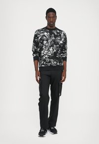 Felpa nera con intricate decorazioni floreali bianche, abbinata a pantaloni cargo neri con tasche laterali. Modello in piedi su uno sfondo bianco.