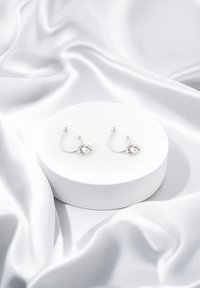 Silberne Creolen mit weißen, sphärischen Akzenten, präsentiert auf einem runden weißen Podest vor einem glatten, weißen Satin-Hintergrund.