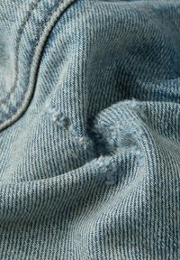 Vaaleansininen farkkukangas, jossa on teksturoitu kudonta, hieman rispaantunut reuna ja pehmeä, rypistynyt ulkonäkö.