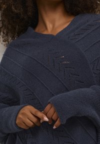 Marinblå stickad tröja med V-ringning, chevront mönster och lösa ärmar med ribbade muddar, som visar en texturerad tyg.