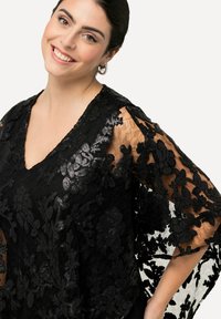 Top de encaje floral negro con escote en V, mangas transparentes y detalles intrincados de lentejuelas. La tela tiene una apariencia delicada y texturizada.