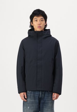 Svart vanntett jakke med hette; har minimalistisk design, lange ermer og en jevn tekstur; båret over slitt denimjeans.
