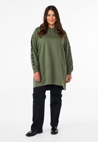 Felpa verde con cappuccio e maniche lunghe, con la scritta "STAY WARM" sulla manica sinistra; abbinata a pantaloni neri e scarpe nere lucide.