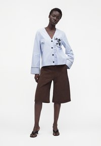 Femme portant un chemisier boutonné rayé bleu clair avec broderie noire et un short marron foncé longueur genou avec des sandales noires à talons.