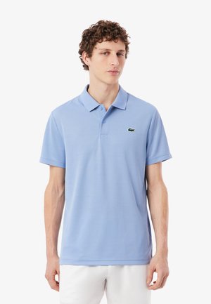 Hombre con camisa polo de manga corta color azul claro con un pequeño logo de cocodrilo verde, combinado con pantalones blancos, de pie frente a un fondo blanco.
