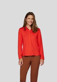 Red long-sleeve button-up shirt with a collar, smooth texture, and slightly loose fit, paired with brown pants.