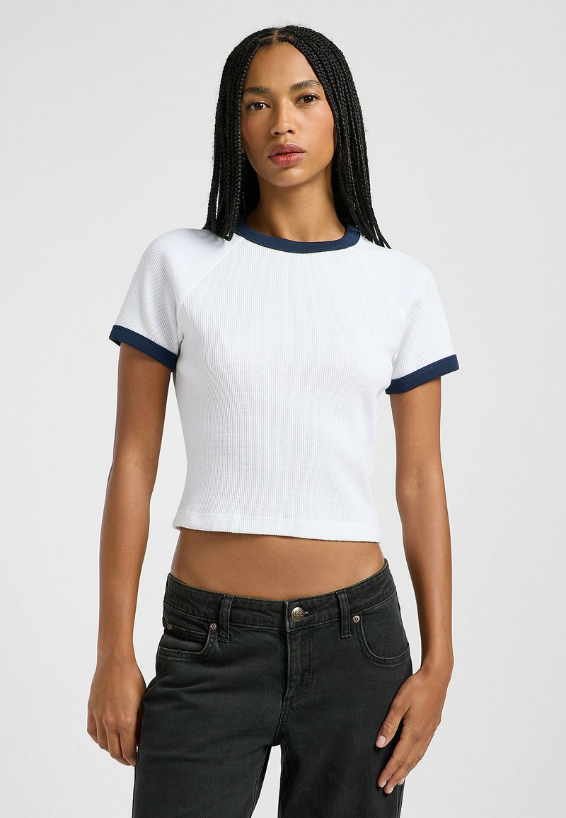 Witte geribbelde crop top met korte mouwen en marineblauwe accenten op de ronde halslijn en mouwranden, gecombineerd met zwarte high-waisted jeans.