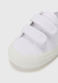 Zapatilla de lona blanca con doble tira de Velcro, puntera redondeada y suela de goma texturizada. Presenta una superficie de tela suave y detalles cosidos.