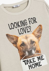 Beige sweatshirt met een grote afbeelding van het gezicht van een hond met de tekst "OP ZOEK NAAR LIEFDE?" erboven en "NEEM ME MEE NAAR HUIS" eronder.