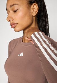 Langermet treningsgenser i myk taupe med hvite stripeaksenter, featuring en rund hals og subtilt logo. Glatt tekstur.