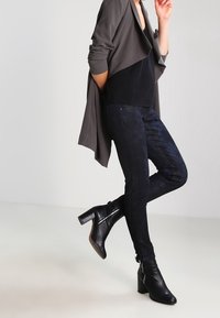 Cardigan gris drapeado sobre una blusa negra, combinado con jeans oscuros de estampado floral y bufandas de tacón negro. Textura suave y diseño ajustado.