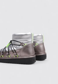 Baskets montantes argentées et gris mat avec des lacets noirs et des accents vert néon. La semelle texturée affiche une marque sur le côté.