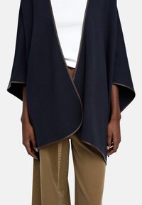 Granatowy wełniany poncho o kroju zawiązywanym, z kontrastującymi brązowymi wykończeniami i szerokimi rękawami, noszone na białym topie i beżowych spodniach.