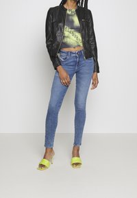 Czarna skórzana kurtka typu bomber, krótki top z grafiką, niebieskie obcisłe dżinsy oraz jaskrawożółte mule na obcasie z plecionym wzorem.