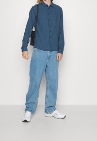 Persoon in een blauw geruit overhemd, losse lichtblauwe spijkerbroek, witte sneakers en met een kleine zwarte schoudertas voor een effen achtergrond.