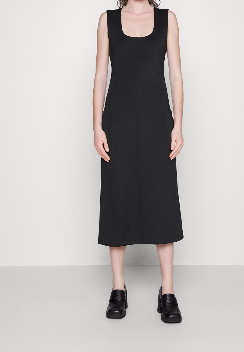 Robe midi sans manches noire avec un décolleté carré, fabriquée en tissu lisse. Présente une coupe droite et tombe jusqu'au mollet. Associée à des chaussures à plateforme noires.