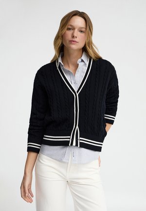 Jeune femme portant un cardigan noir en maille torsadée avec bordure blanche par-dessus une chemise rayée et un pantalon blanc, sur un fond uni.