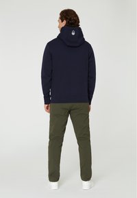 Sudadera azul marino con bolsillo tipo canguro delantero, capucha con cordón y logo en la parte posterior. Combinada con pantalones verde oliva y zapatillas blancas.