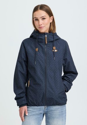 Jonge vrouw met een marineblauwe jas met een capuchon en witte polka dots, met een ritssluiting aan de voorkant, en lichtblauwe spijkerbroek, staand met één hand in de zak.