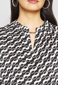 Femme portant un chemisier noir avec un motif chaîne blanc, un détail barre argentée au niveau de l'encolure et de grandes boucles d'oreilles argentées en forme d'anneau.