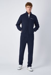 Navy zip-up treningsdress i myk fabric, med høy krage, sidelommer og logo detaljer på brystet. Kombinert med matchende bukser.