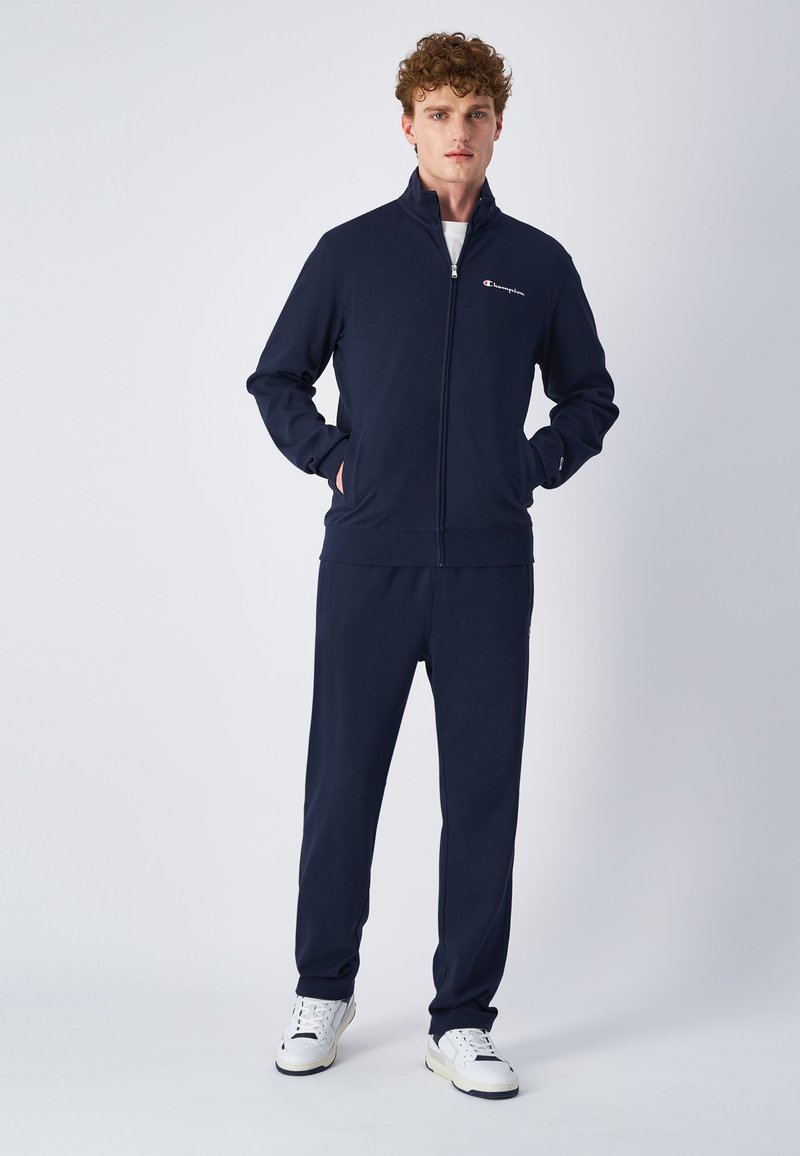 Navy zip-up treningsdress i myk fabric, med høy krage, sidelommer og logo detaljer på brystet. Kombinert med matchende bukser.