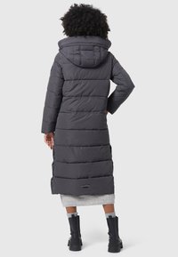 Long puffer coat in dark grey, quilted design with a hood, featuring a logo patch near the hem and long sleeves. Worn over a light grey skirt.
