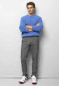 Junger Mann steht mit verschränkten Armen, trägt einen blauen Pullover, graue Hose und weiße Sneaker mit roten Akzenten vor einem schlichten Hintergrund.