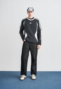 Fekete Adidas póló fehér díszítéssel és csíkokkal, hosszú ujjú ingre rétegezve. Sötét farmernadrággal és fehér tornacipővel viselve.