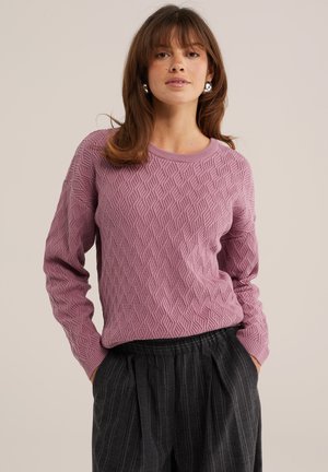 Mujer con un suéter morado texturizado y pantalones oscuros de rayas amplios, con las manos en los bolsillos, sobre un fondo liso.
