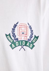 Balta T-kreklu ar krāsainu Adidas logotipu. Dizains ietver sarkanu, zilu un zaļu motīvu ar lauru lapām un tekstu.