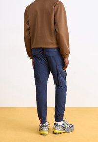 Brązowy sweter z prążkowanymi ściągaczami, w połączeniu z granatowymi spodniami cargo z kieszeniami po bokach i z tyłu. Szaro-żółte sneakersy dopełniają stylizację.