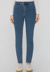 Džíny v modré barvě s vysok ým pasem a slim fit, obsahující pět kapes, poutka na opasek a kontrastní švy. Nosené s bílými teniskami.