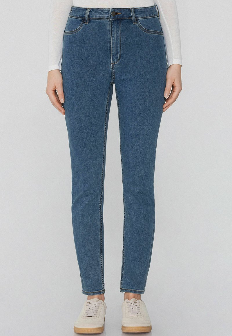 Džíny v modré barvě s vysokým pasem a slim fit, obsahující pět kapes, poutka na opasek a kontrastní švy. Nosené s bílými teniskami.