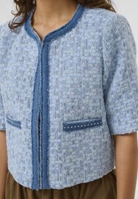 Hellblaue Tweedjacke mit kurzen Ärmeln, ausgefransten Kanten, blauen Akzenten und dekorativen Taschen, die entlang der Ränder mit Nieten verziert sind.