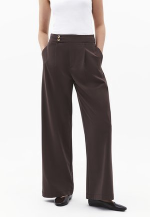 Kobieta w ciemnobrązowych spodniach o szerokich nogawkach z detalem guzików w talii, w połączeniu z czarnymi balerinami i białym topem bez rękawów. Ręce w kieszeniach.