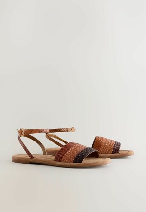 Par niti ravne smeđe tkanih kožnih sandala s remenima oko članka i kopčama na svijetlo neutralnoj pozadini.