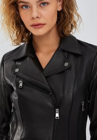 Giacca da motociclista in pelle nera con colletto a falde, zip prominenti e diverse tasche con zip, caratterizzata da una texture liscia e un design aderente.