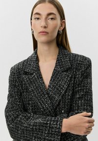 Melnbalts raksts ar struktūrētu audumu, pla�šām atlokām un dubultā pogu dizains. Modele ar rokām sakrustotām, neitrālā fonā.