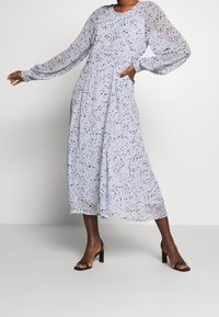 Hellblaues langärmliges Kleid mit verstreuten schwarzen Punkten, gerafftem Taillenbund und fließender Silhouette. Kombiniert mit schwarzen Absatzsandalen.