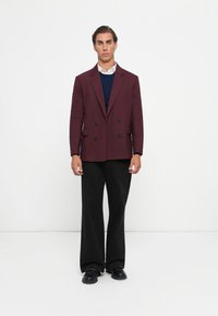 Mand står mod en hvid baggrund iført en kastanjefarvet dobbeltbrystet blazer, en marineblå sweater, en hvid skjorte, sorte bukser og sorte sko.