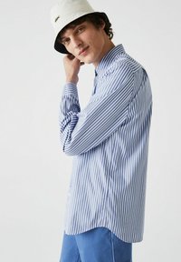 Camisa de botones a rayas en tela azul claro y blanca, con mangas largas y dobladillo redondeado. Llevada con un sombrero tipo bucket blanco.